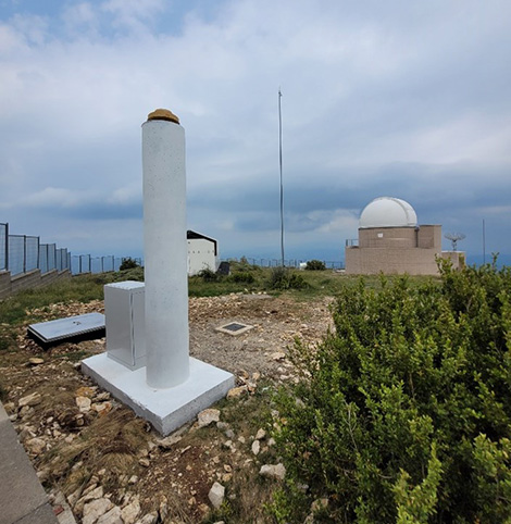 Pilar geodèsic d'una estació de referència GNSS, amb l'armari d'equipament a la base i, al fons, la cúpula d'un observatori astronòmic.