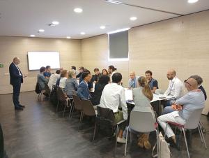 Several people sitting around a table participating in a working session. A presentation is projected on the screen in the background, while on the table there is a work panel, markers and notes used during the dynamic.