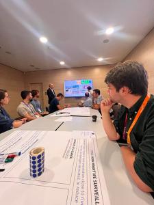 Room with people sitting around two long tables participating in a working session. In the background is a presentation screen and a person standing next to the screen.