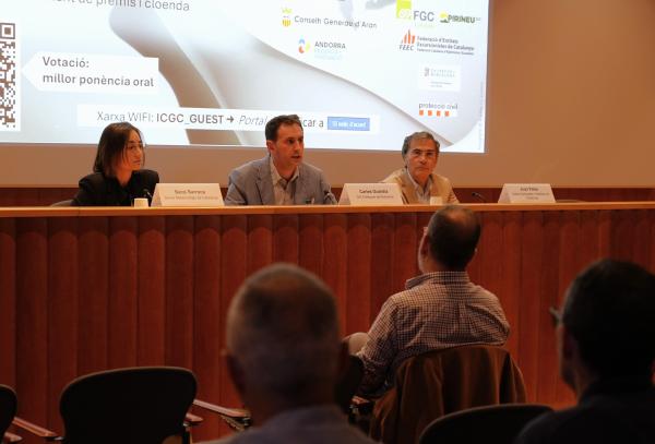 Tres persones, una dona i dos homes, seuen darrere d’una taula de fusta amb cartells identificatius davant seu. Darrere, una pantalla projecta una diapositiva amb logotips d’entitats organitzadores i la informació de la xarxa wifi. En primer terme, es veuen d’esquena alguns assistents.