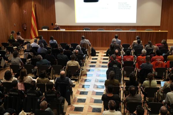 Vista elevada de la sala d’actes plena de públic assegut, amb alguns assistents amb uniforme. Al fons, un ponent assegut darrere d’una taula i una pantalla amb una diapositiva projectada. A l’esquerra hi ha una bandera de Catalunya.