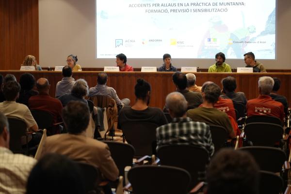 Sala d’actes amb públic assegut, vist d’esquena, davant d’una taula amb sis persones ponents. Darrere seu, una pantalla projecta el títol de la taula rodona i els logotips de les entitats organitzadores.