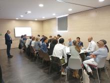 Several people sitting around a table participating in a working session. A presentation is projected on the screen in the background, while on the table there is a work panel, markers and notes used during the dynamic.