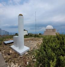 Pilar geodèsic d'una estació de referència GNSS, amb l'armari d'equipament a la base i, al fons, la cúpula d'un observatori astronòmic.